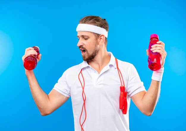 Selbstbewusster junger, gutaussehender, sportlicher Mann mit Stirnband und Armbändern, der Wasserflaschen hält, die mit Springseil um den Hals isoliert auf blauer Wand nach unten schauen