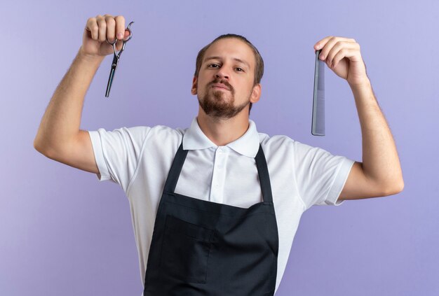 Selbstbewusster junger hübscher Friseur, der Uniform hält, die Schere und Kamm hält, lokalisiert auf lila