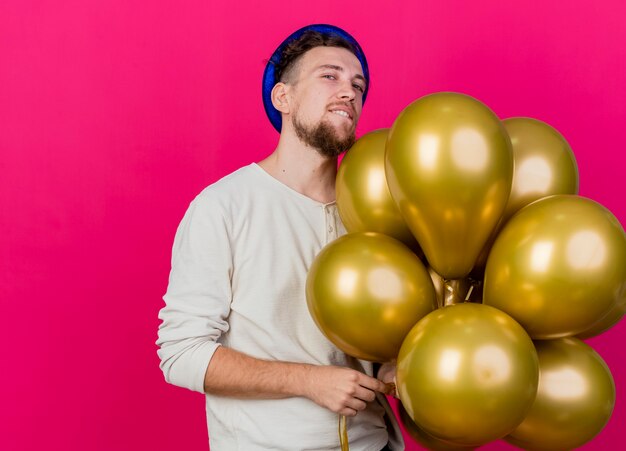 Selbstbewusster junger hübscher Party-Typ, der Partyhut hält, der Ballons hält, die auf vordere beißende Lippe lokalisiert auf purpurroter Wand betrachten