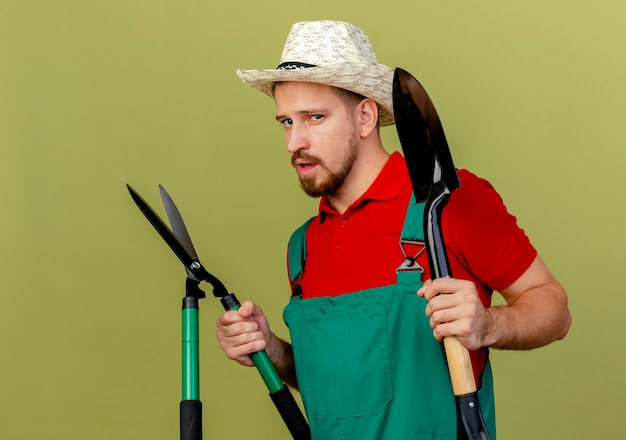 Selbstbewusster junger hübscher slawischer Gärtner in der Uniform und im Hut, die in der Profilansicht stehen, die Scheren und Spaten lokalisiert auf olivgrüner Wand hält