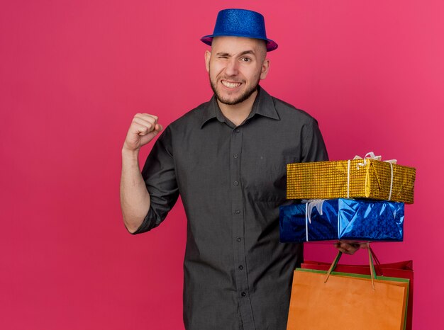 Selbstbewusster junger hübscher slawischer Party-Typ, der Partyhut hält, der Geschenkverpackungen und und Papiertüten hält