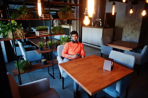 Selbstbewusster junger Inder in schicker Freizeitkleidung wie orangefarbenem Pullover, der im Café sitzt und sein Handy benutzt