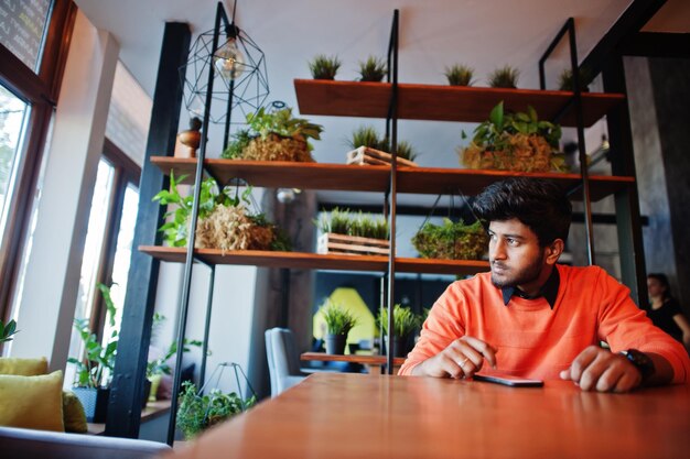 Selbstbewusster junger Inder in schicker Freizeitkleidung wie orangefarbenem Pullover, der im Café sitzt und sein Handy benutzt