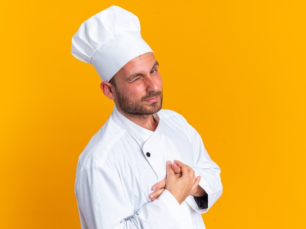Selbstbewusster junger kaukasischer männlicher Koch in Kochuniform und Mütze, der in der Profilansicht steht und die Hände auf der Brust hält und in die Kamera schaut, die isoliert auf oranger Wand zwinkert