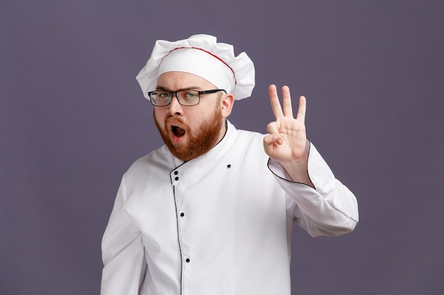 Selbstbewusster junger Koch mit Brille, Uniform und Mütze, der in die Kamera blickt und ein Ok-Zeichen auf violettem Hintergrund zeigt