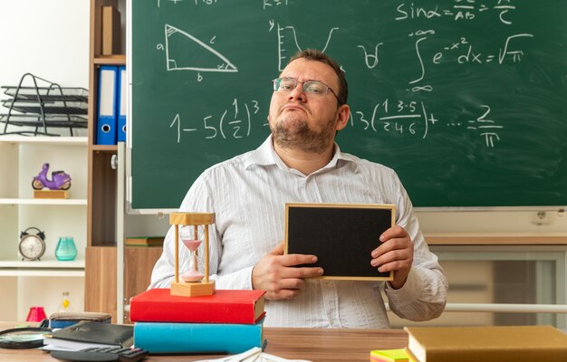 selbstbewusster junger Lehrer mit Brille, der am Schreibtisch mit Schulmaterial im Klassenzimmer sitzt und eine Mini-Tafel zeigt, die nach vorne schaut