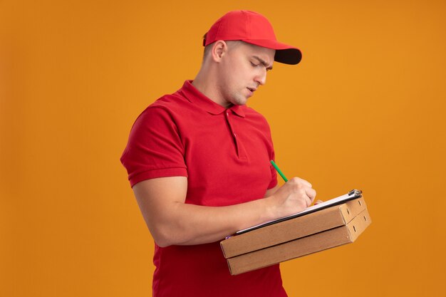 Selbstbewusster junger Lieferbote, der Uniform mit Kappe hält, die Pizzaschachteln hält und etwas auf Zwischenablage schreibt, die auf orange Wand lokalisiert wird