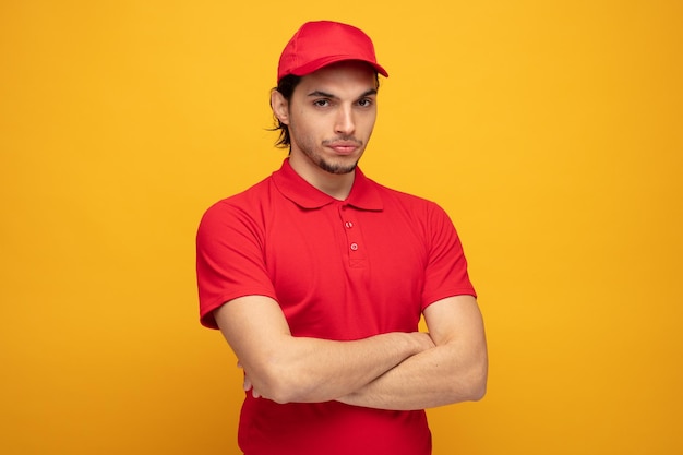 Selbstbewusster junger Lieferbote in Uniform und Mütze, der in die Kamera blickt, während er die Arme isoliert auf gelbem Hintergrund verschränkt hält