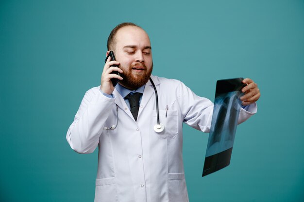 Selbstbewusster junger männlicher Arzt mit Arztkittel und Stethoskop um den Hals, der Röntgenaufnahmen hält und betrachtet, während er telefoniert, isoliert auf blauem Hintergrund