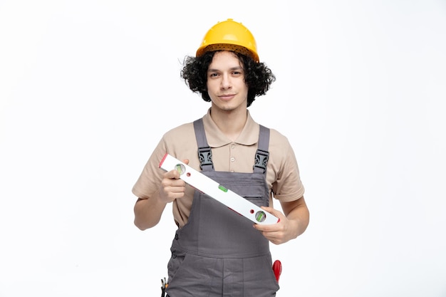 Selbstbewusster junger männlicher Bauarbeiter in Uniform und Schutzhelm mit Wasserwaage, der mit Bauinstrumenten in der Tasche isoliert auf weißem Hintergrund in die Kamera blickt