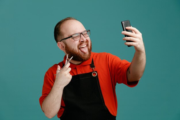 Selbstbewusster junger männlicher Friseur mit Brille, rotem Hemd und Friseurschürze mit Schere und Zunge, die Selfie mit Handy isoliert auf blauem Hintergrund macht