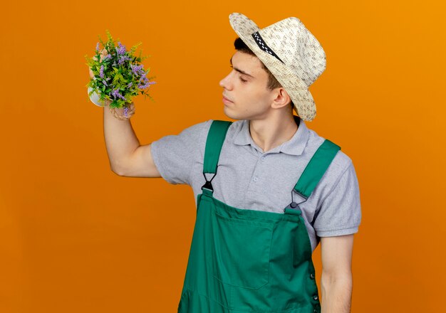 Selbstbewusster junger männlicher Gärtner, der Gartenhut trägt, hält und schaut auf Blumentopf