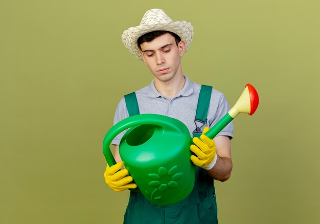 Selbstbewusster junger männlicher Gärtner, der Gartenhut und Handschuhe trägt, hält und schaut auf Gießkanne