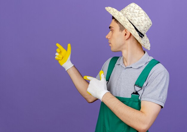 Selbstbewusster junger männlicher Gärtner, der Gartenhut und Handschuhe trägt, schaut und zeigt nach hinten