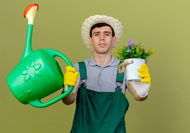 Selbstbewusster junger männlicher Gärtner mit Gartenhut und Handschuhen hält Gießkanne