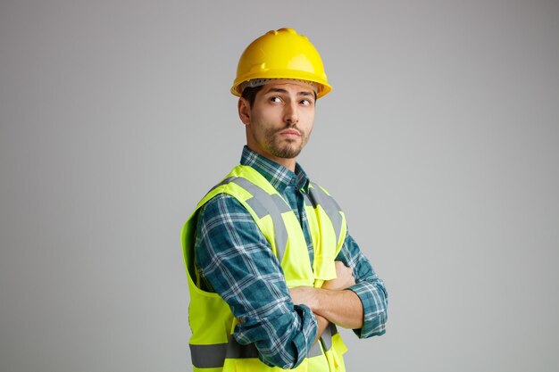 Selbstbewusster junger männlicher Ingenieur mit Schutzhelm und Uniform, der in der Profilansicht steht und zur Seite schaut, während er die Arme verschränkt hält, isoliert auf weißem Hintergrund mit Kopierraum