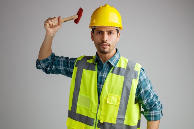 Selbstbewusster junger männlicher Ingenieur mit Schutzhelm und Uniform, der in die Kamera blickt, die den Hammer isoliert auf weißem Hintergrund hebt