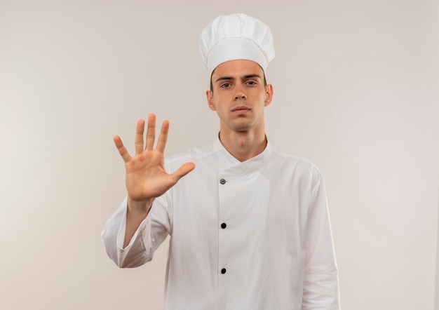 selbstbewusster junger männlicher Koch, der Kochuniform trägt, die Stoppgeste auf isolierter weißer Wand zeigt