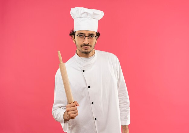 Selbstbewusster junger männlicher Koch, der Kochuniform und Gläser hält, die Nudelholz halten
