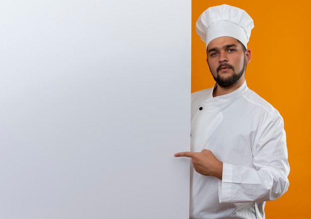 Selbstbewusster junger männlicher Koch in Kochuniform, der dahinter steht und auf die weiße Wand zeigt, die auf einer orangefarbenen Wand mit Kopienraum isoliert ist?