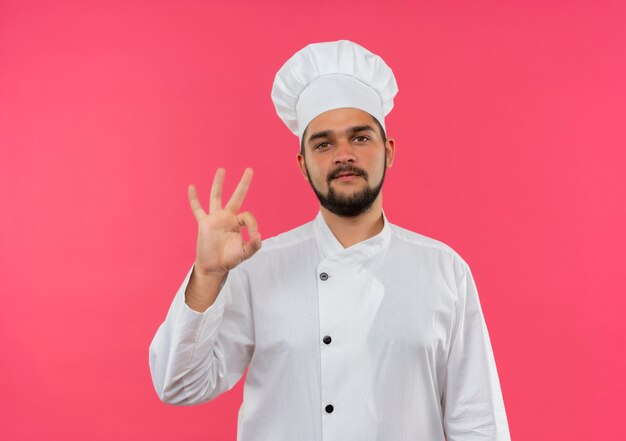 Selbstbewusster junger männlicher Koch in Kochuniform, der das OK-Zeichen einzeln auf rosa Wand mit Kopienraum tut