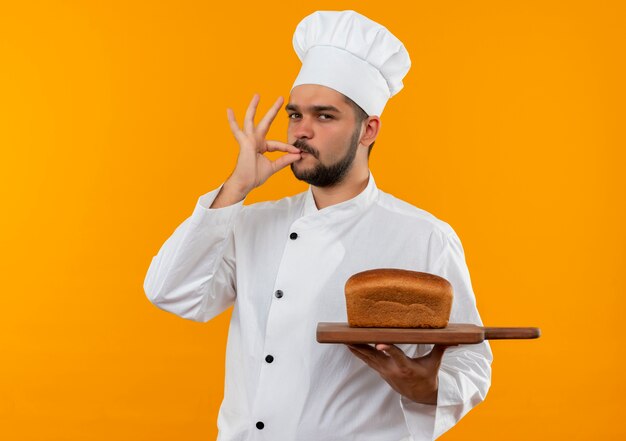 Selbstbewusster junger männlicher Koch in Kochuniform, der ein Schneidebrett mit Brot darauf hält und eine leckere Geste einzeln auf der orangefarbenen Wand macht