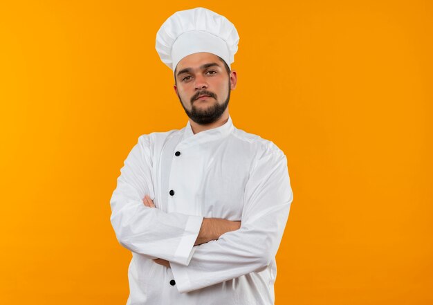Selbstbewusster junger männlicher Koch in Kochuniform, der mit geschlossener Haltung isoliert auf oranger Wand mit Kopierraum steht standing