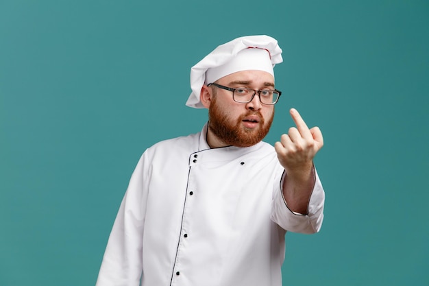 Selbstbewusster junger männlicher koch mit brille, uniform und mütze, der auf die kamera blickt und eine geste isoliert auf blauem hintergrund macht