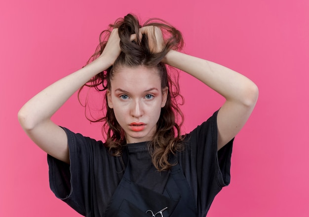 Selbstbewusster junger slawischer weiblicher friseur, der uniform trägt, die ihr haar ergreifend betrachtet kamera lokalisiert auf rosa hintergrund