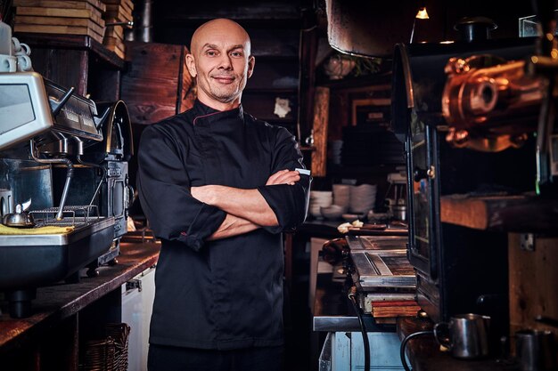 Selbstbewusster Koch in Uniform, der mit verschränkten Armen posiert und in einer Restaurantküche auf eine Kamera blickt.