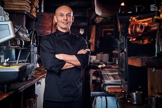 Selbstbewusster Koch in Uniform, der mit verschränkten Armen posiert und in einer Restaurantküche auf eine Kamera blickt.