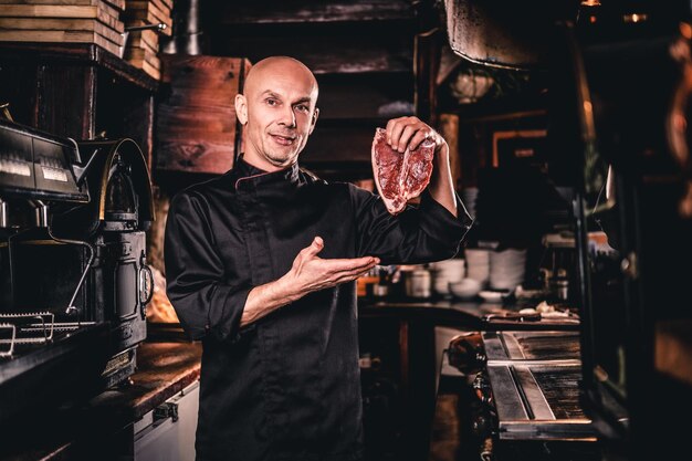 Selbstbewusster Koch in Uniform, der vor dem Kochen ein frisches Steak präsentiert und eine Kamera in einer Restaurantküche anschaut.