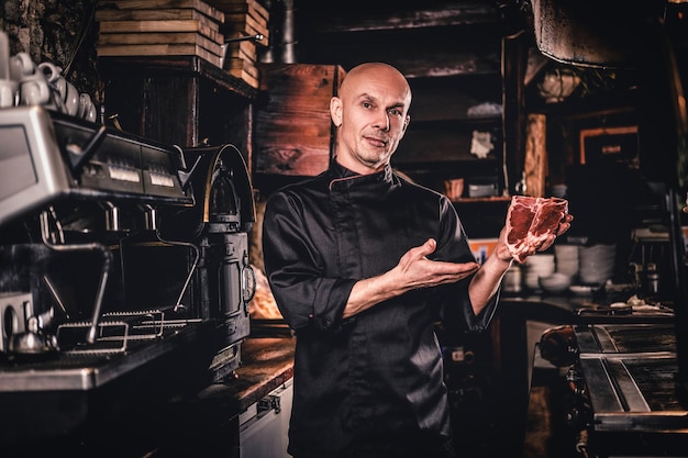 Selbstbewusster Koch in Uniform, der vor dem Kochen ein frisches Steak präsentiert und eine Kamera in einer Restaurantküche anschaut.
