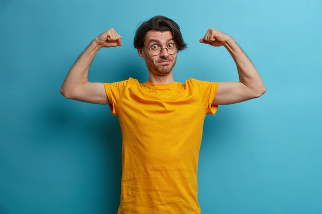Selbstbewusster, kraftvoller Mann hebt die Arme und zeigt Muskeln, zeigt das Ergebnis eines regelmäßigen Trainings, trägt ein gelbes T-Shirt und eine Brille, führt einen aktiven, gesunden Lebensstil und ist sehr stark