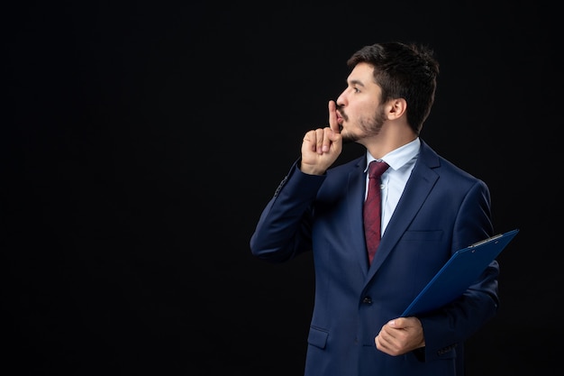 Selbstbewusster männlicher Büroangestellter im Anzug, der Dokumente hält und an isolierter dunkler Wand eine Stille-Geste macht