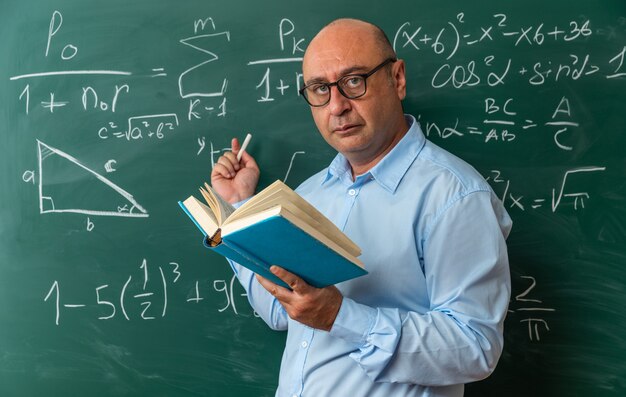 Selbstbewusster männlicher Lehrer mittleren Alters mit Brille, der vor der Tafel steht und ein Buch hält