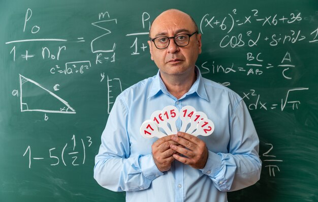 Selbstbewusster männlicher Lehrer mittleren Alters mit Brille, der vor der Tafel steht und Nummernfächer hält