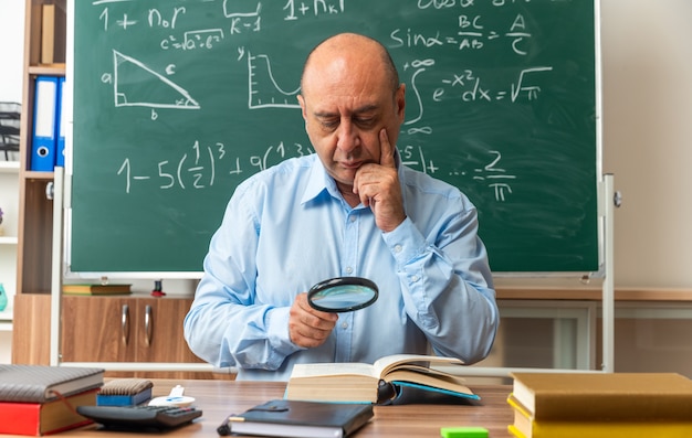 Selbstbewusster männlicher lehrer mittleren alters sitzt am tisch mit schulmaterial und liest ein buch mit lupe, das die hand ins klassenzimmer legt