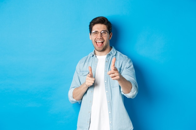 Selbstbewusster mann, der glückwunsch sagt, zwinkert und auf dich zeigt, erfreut über blauem hintergrund steht und lächelt
