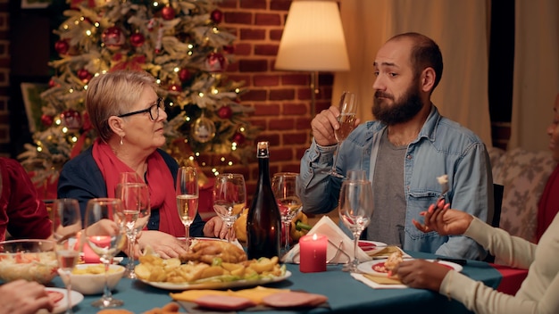 Selbstbewusster Mann, der mit positiver Seniorin spricht, während er beim Weihnachtsessen Sekt trinkt. Fröhliche gemischtrassige Familienmitglieder feiern das Winterfest mit traditionellem Essen und Champagner.