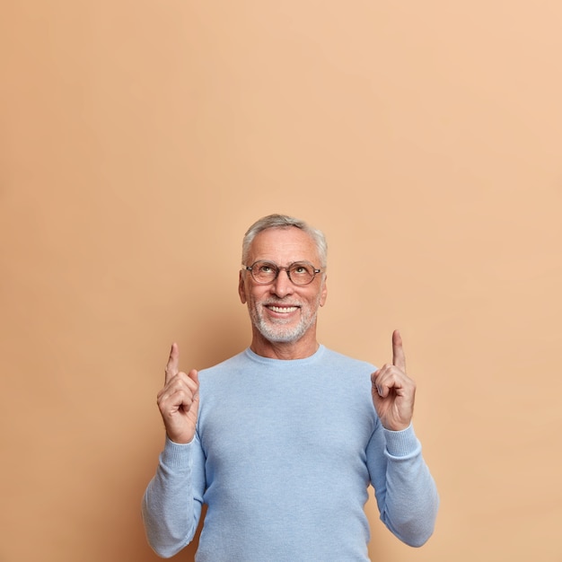 Selbstbewusster positiver reifer Mann zeigt oben auf Leerzeichen und zeigt Werbung über dem Kopf, gekleidet in einen lässigen blauen Pullover, der über einer braunen Wand isoliert ist