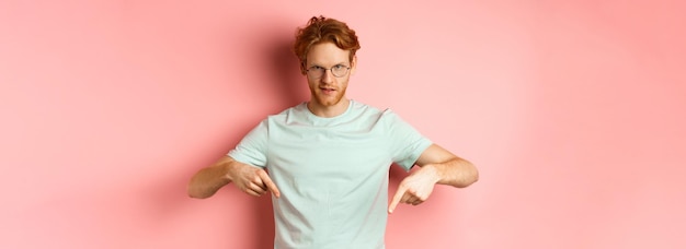 Kostenloses Foto selbstbewusster rothaariger mann mit brille, der mit dem finger nach unten zeigt und gewagt mit selbstgefälligem gesicht in die kamera starrt