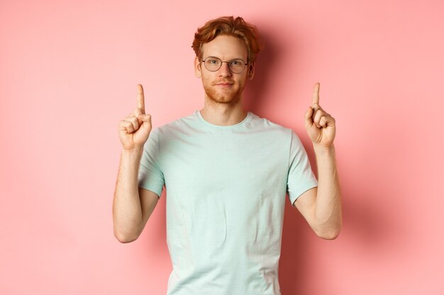 Selbstbewusster und zufriedener junger Mann mit roten Haaren mit Brille und T-Shirt, der mit den Fingern nach oben zeigt und lächelt.