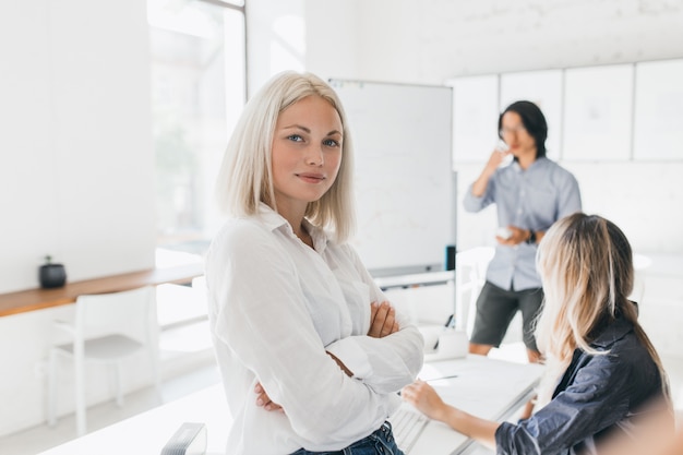 Selbstbewusstes blondes Mädchen in der Bluse, die mit verschränkten Armen im Büro mit großem Flipchart steht