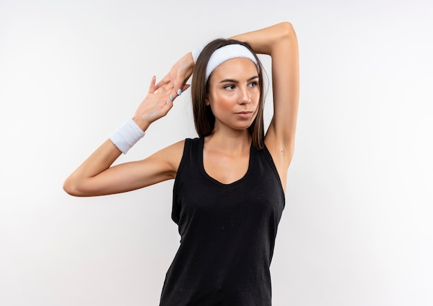 Selbstbewusstes hübsches sportliches Mädchen mit Stirnband und Armband, das den Arm auf den Kopf legt und die Hände zusammenhält und die Seite isoliert auf weißer Wand betrachtet