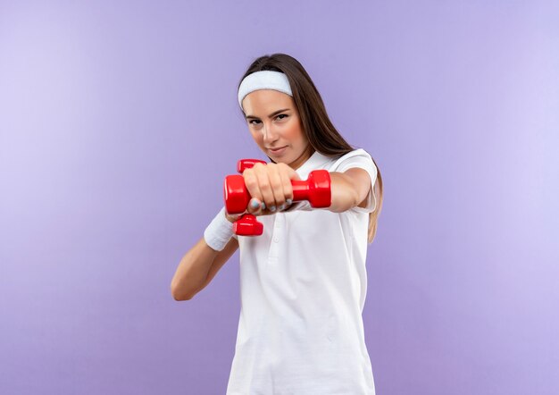 Selbstbewusstes hübsches sportliches Mädchen mit Stirnband und Armband, das Hantel isoliert auf lila Wand ausstreckt