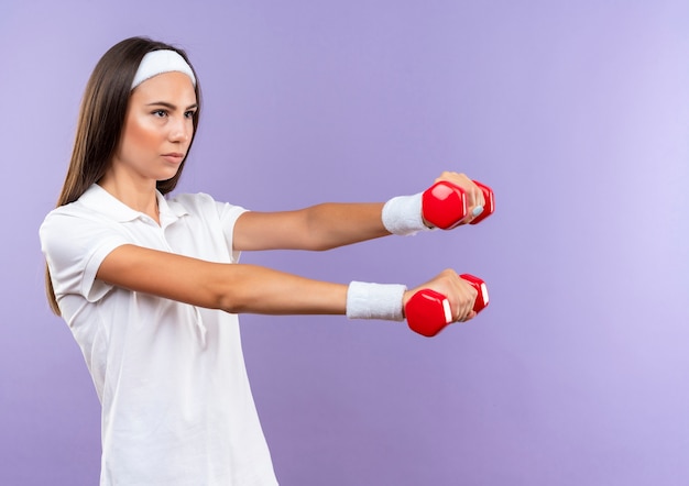 Selbstbewusstes hübsches sportliches Mädchen mit Stirnband und Armband, das Hanteln isoliert auf lila Wand mit Kopienraum hält holding