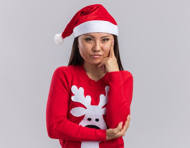 Selbstbewusstes junges asiatisches Mädchen mit Weihnachtsmütze mit Pullover, das den Finger auf die Wange legt, isoliert auf weißem Hintergrund