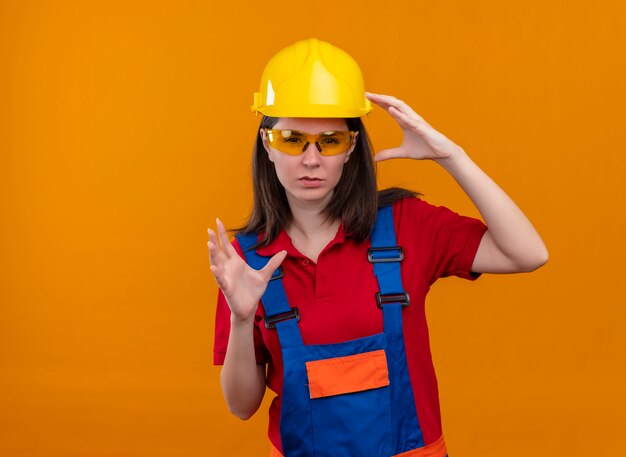 Selbstbewusstes junges Baumeistermädchen mit Sicherheitsbrille hält Hände hoch auf lokalisiertem orangefarbenem Hintergrund mit Kopienraum