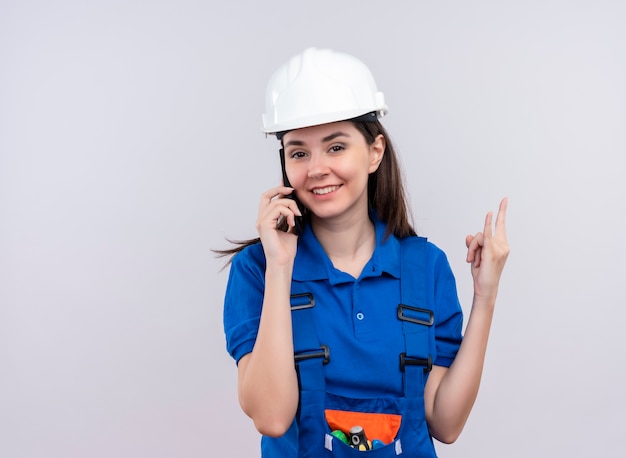 Selbstbewusstes junges Baumeistermädchen mit weißem Schutzhelm und blauer Uniform spricht am Telefon und gestikuliert Zeichen der Hörner auf lokalisiertem weißem Hintergrund mit Kopienraum